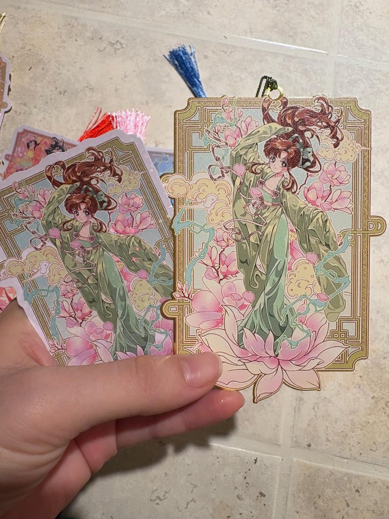 May include: Bookmarks featuring a detailed illustration of a woman in a flowing green gown, surrounded by pink flowers and clouds. The bookmarks have a gold border and a tassel attached to the top.