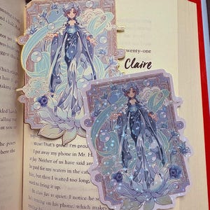 May include: Two decorative bookmarks featuring a detailed illustration of a woman in a flowing blue gown, surrounded by bubbles and floral accents. The bookmarks are set against the pages of a book. The word "Claire" is visible on the page.