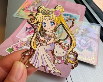 May include: A gold-colored enamel pin featuring a cartoon character with blonde hair, a crown, and a long dress, holding a staff. A Hello Kitty character is seated beside her. Pink packaging and stickers are in the background.