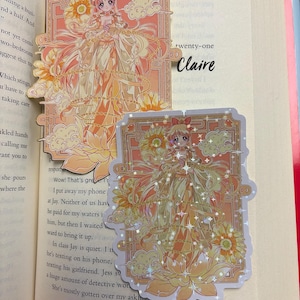 May include: A bookmark and sticker featuring a detailed anime-style illustration of a character in a flowing gown, with floral and cloud motifs in shades of orange and yellow. The bookmark has a gold chain.