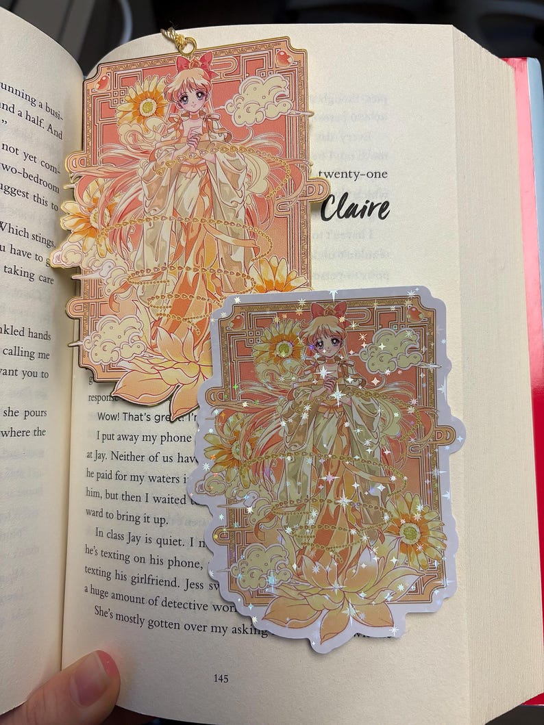 May include: A bookmark and sticker featuring a detailed anime-style illustration of a character in a flowing gown, with floral and cloud motifs in shades of orange and yellow. The bookmark has a gold chain.