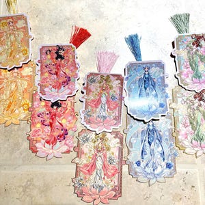 May include: A collection of decorative bookmarks featuring illustrations of women in ornate dresses, set against floral and celestial backgrounds. Each bookmark has a colorful tassel. The color palette includes pink, blue, gold, and orange.