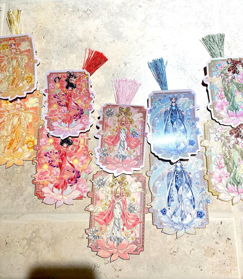May include: A collection of decorative bookmarks featuring illustrations of women in ornate dresses, set against floral and celestial backgrounds. Each bookmark has a colorful tassel. The color palette includes pink, blue, gold, and orange.