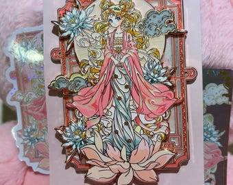 May include: A pink and gold enamel pin featuring a woman in a flowing gown, surrounded by lotus flowers and clouds. The pin is set against a pink background with a matching sticker and a pink plush toy.
