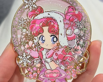 May include: A circular enamel pin featuring a cartoon character with pink hair and a winter outfit. The pin has a gold border with snowflake and floral designs. The character is wearing a white-trimmed hat and a pink dress.