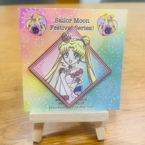 Könnte beinhalten: Eine rosa und blaue Emaille-Anstecknadel mit Sailor Moon in einem weißen Kleid mit rosa Blumen und einer Krone. Die Anstecknadel hat eine Diamantenform mit einem goldenen Rand und befindet sich auf einer Karte mit einem Regenbogenhintergrund. Die Karte trägt den Text "Sailor Moon Festival Series!" und die Adresse "princessmoonpins.etsy.com".