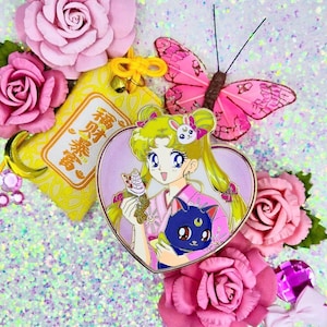 Könnte beinhalten: Eine rosa herzförmige Emaille-Anstecknadel mit einer Comicfigur mit blonden Haaren, einem rosa Kleid und einer schwarzen Katze. Die Figur hält einen japanischen Nachtisch und ein kleines weißes Kaninchen. Die Anstecknadel ist von rosa Blumen und einer gelben Stofftasche mit japanischen Schriftzeichen umgeben.