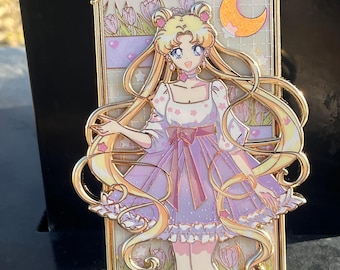 May include: A gold-toned enamel pin featuring a cartoon character with long blonde hair, wearing a purple dress with white polka dots and a pink bow. The character is surrounded by a gold frame and a crescent moon.