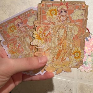 May include: Two anime-style bookmarks featuring a character in a flowing yellow dress with a bow in their hair. The bookmarks have a floral and cloud design with gold accents. One bookmark has a blue tassel.