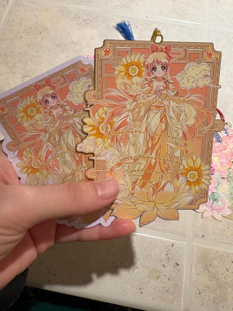 May include: Two anime-style bookmarks featuring a character in a flowing yellow dress with a bow in their hair. The bookmarks have a floral and cloud design with gold accents. One bookmark has a blue tassel.
