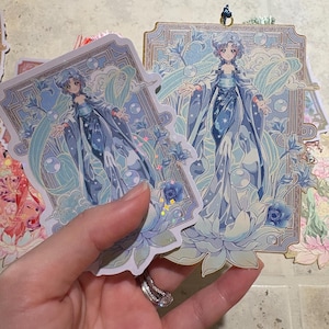 May include: Two stickers featuring a detailed illustration of a woman in a flowing blue gown, surrounded by floral and water-themed elements. The stickers have a light blue and gold color scheme, with a decorative border.