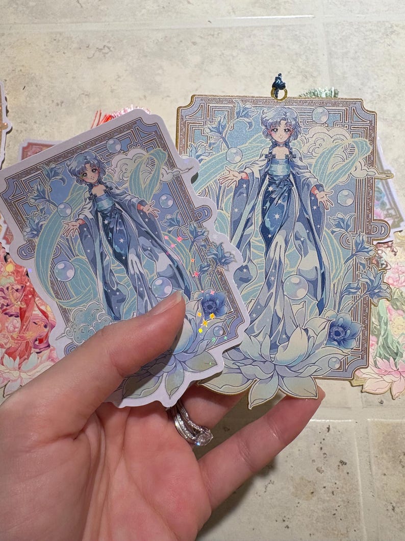 May include: Two stickers featuring a detailed illustration of a woman in a flowing blue gown, surrounded by floral and water-themed elements. The stickers have a light blue and gold color scheme, with a decorative border.