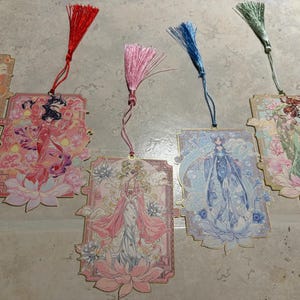 May include: A collection of decorative bookmarks featuring illustrations of women in flowing gowns, with floral and celestial motifs. Each bookmark has a tassel in a different color, including red, pink, blue, and green.