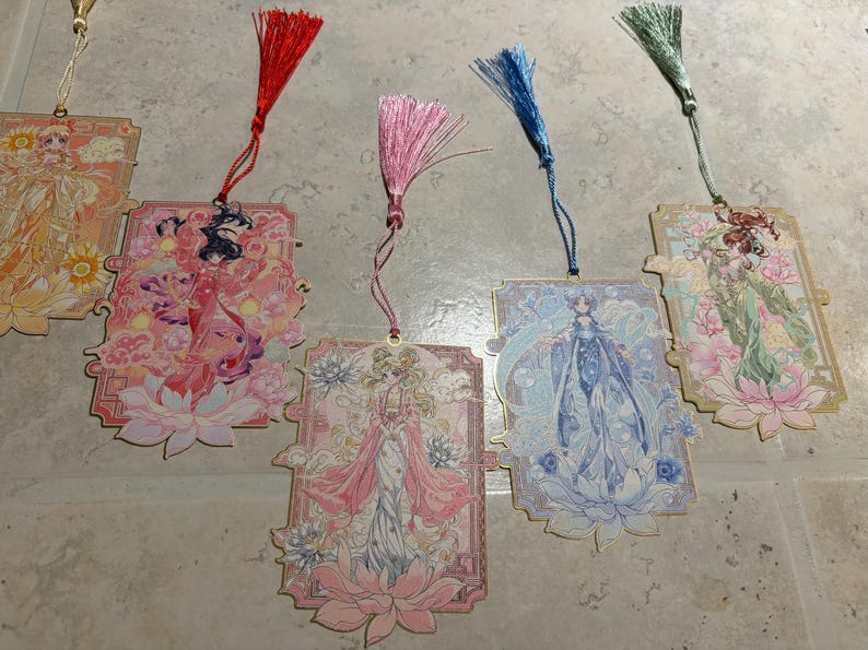 May include: A collection of decorative bookmarks featuring illustrations of women in flowing gowns, with floral and celestial motifs. Each bookmark has a tassel in a different color, including red, pink, blue, and green.