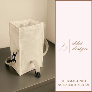 May include: Cream-coloured thermal-lined insulated lunch bag with a rectangular shape and a handle. The bag has a buckle closure and a strap. The text "diloi design" is visible on the right side of the image. The words "THERMAL-LINED INSULATED LUNCH BAG" are also visible.