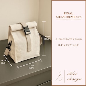 May include: A cream-coloured canvas lunch bag with a roll-top closure and a black buckle. The bag has a shoulder strap and measures 21cm x 32cm x 16cm. The bag is labeled with the text "FINAL MEASUREMENTS" and "diloi design".