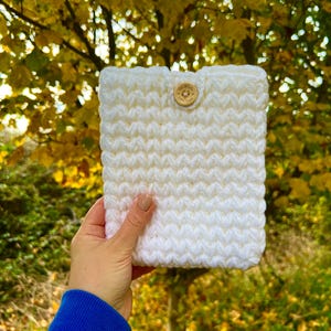 以下が含まれることがあります： 白の、かぎ針編みで作られたタブレットケース。テクスチャード加工のパターンと木製のボタン留めが特徴です。秋の葉を背景に、手作りの品質とニュートラルな色合いを際立たせています。