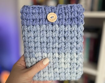 May include: A handmade, blue crochet book sleeve with a button closure. The sleeve features a gradient of blue hues, transitioning from a darker shade at the top to a lighter shade at the bottom. The wooden button is engraved with the words "handmade with love."