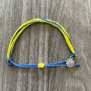 May include: A blue and yellow friendship bracelet with a silver sunflower charm. The bracelet is made of braided string and has a knot at the end.