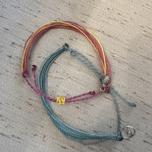 May include: Two colorful friendship bracelets with a silver charm on one of the bracelets. The bracelets are made of woven string in shades of blue, pink, yellow, and red.