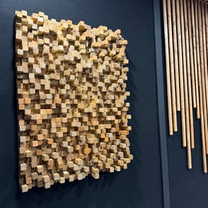 May include: A wall-mounted sound diffuser made of wood. The diffuser is made up of many small, square blocks of wood that are arranged in a random pattern. The diffuser is a light brown color and is mounted on a black wall.