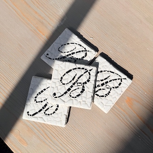Puede incluir: Cuatro posavasos cuadrados blancos con un diseño de mosaico. Cada posavasos presenta la letra negra "B" en escritura cursiva. Los posavasos están colocados sobre una superficie de madera clara.
