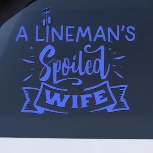 May include: A blue vinyl decal applied to a car window. The decal reads "A Lineman's Spoiled Wife" in a stylized font. The car is white, and the decal is centered on the rear window.