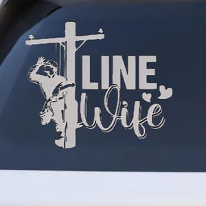 May include: White decal on a car window featuring a lineman on a utility pole and the words "LINE Wife" in a script font. A heart shape is next to the word "Wife".