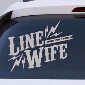 May include: White vinyl car decal with the words "LINE WIFE" in a bold, stylized font, accompanied by lightning bolt graphics. The text "HIGH VOLTAGE" is above the word "WIFE". The decal is affixed to the rear window of a white car.