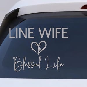 May include: A white car's rear window displays a silver decal. The decal reads "LINE WIFE" above a heart-shaped graphic, and "Blessed Life" in cursive below. The car has a red brake light and a black antenna.