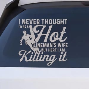 May include: White decal with text: "I NEVER THOUGHT I'D BE A Hot LINEMAN'S WIFE BUT HERE I AM Killing it." The decal features a silhouette of a lineman. The decal is displayed on a car window.