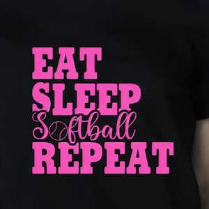 May include: Black t-shirt with the pink text "EAT SLEEP Softball REPEAT". The word "Softball" has a softball graphic in place of the "O". The shirt has short sleeves and a crew neck.