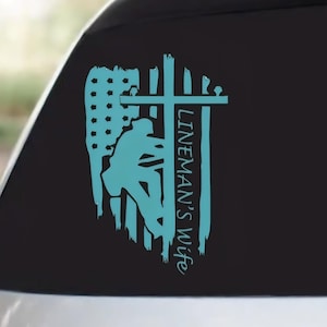 May include: A teal decal featuring a lineman silhouetted against a stylized American flag and cross. The text "LINEMAN'S wife" is vertically aligned on the right side of the design. The decal is applied to a car window.