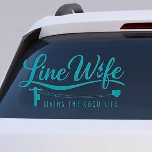 May include: A teal decal on a car window reads "Line Wife" with an illustration of a lineman and fishing poles. Below, the text says "Living the Good Life" with a heart symbol. The decal is on a white car.