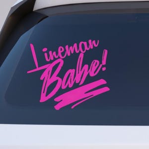 May include: A pink vinyl decal with the words "Lineman Babe!" in a cursive font. The decal is applied to the rear window of a white vehicle. The text is in a bold, handwritten style.