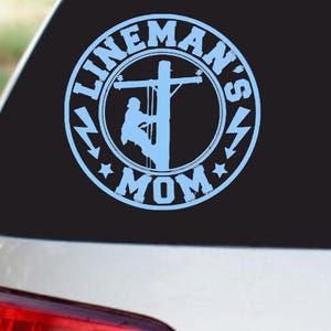 May include: A light blue vinyl decal, featuring a circular design with the text "LINEMAN'S MOM" in a stylized font. The design includes a lineman working on a power pole, surrounded by stars and lightning bolts. The decal is affixed to a car window.