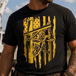 May include: A black short-sleeved t-shirt featuring a yellow graphic print of a stylized American flag with a silhouette of a utility worker climbing a power pole. The design incorporates vertical distressed stripes and stars within the flag pattern.