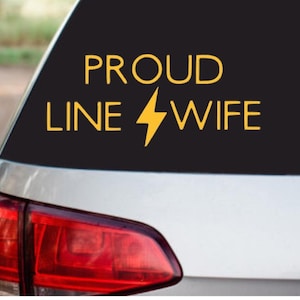 May include: A black car window decal with the words "PROUD LINE WIFE" in gold lettering, with a yellow lightning bolt graphic. The decal is affixed to the rear window of a silver car, with the red taillight visible.