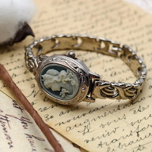 Puede incluir: Un reloj de pulsera plateado con una esfera ovalada que presenta un relieve de camafeo gris de un retrato de perfil. El reloj tiene una pulsera de eslabones met&aacute;licos decorativa con un patr&oacute;n intrincado y una peque&ntilde;a corona lateral.