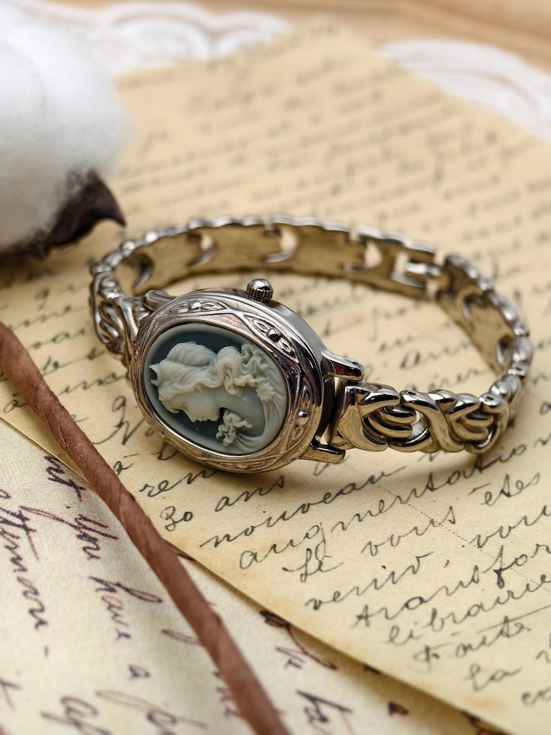Puede incluir: Un reloj de pulsera plateado con una esfera ovalada que presenta un relieve de camafeo gris de un retrato de perfil. El reloj tiene una pulsera de eslabones met&aacute;licos decorativa con un patr&oacute;n intrincado y una peque&ntilde;a corona lateral.