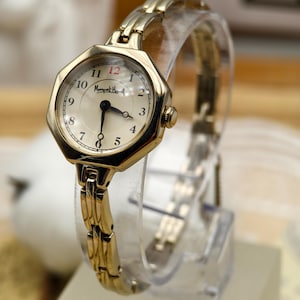 Puede incluir: Un reloj de pulsera dorado con esfera crema y agujas negras. El reloj tiene un bisel octogonal y una correa de eslabones dorados. La esfera muestra la marca "Marquet H." y los números del 1 al 12.