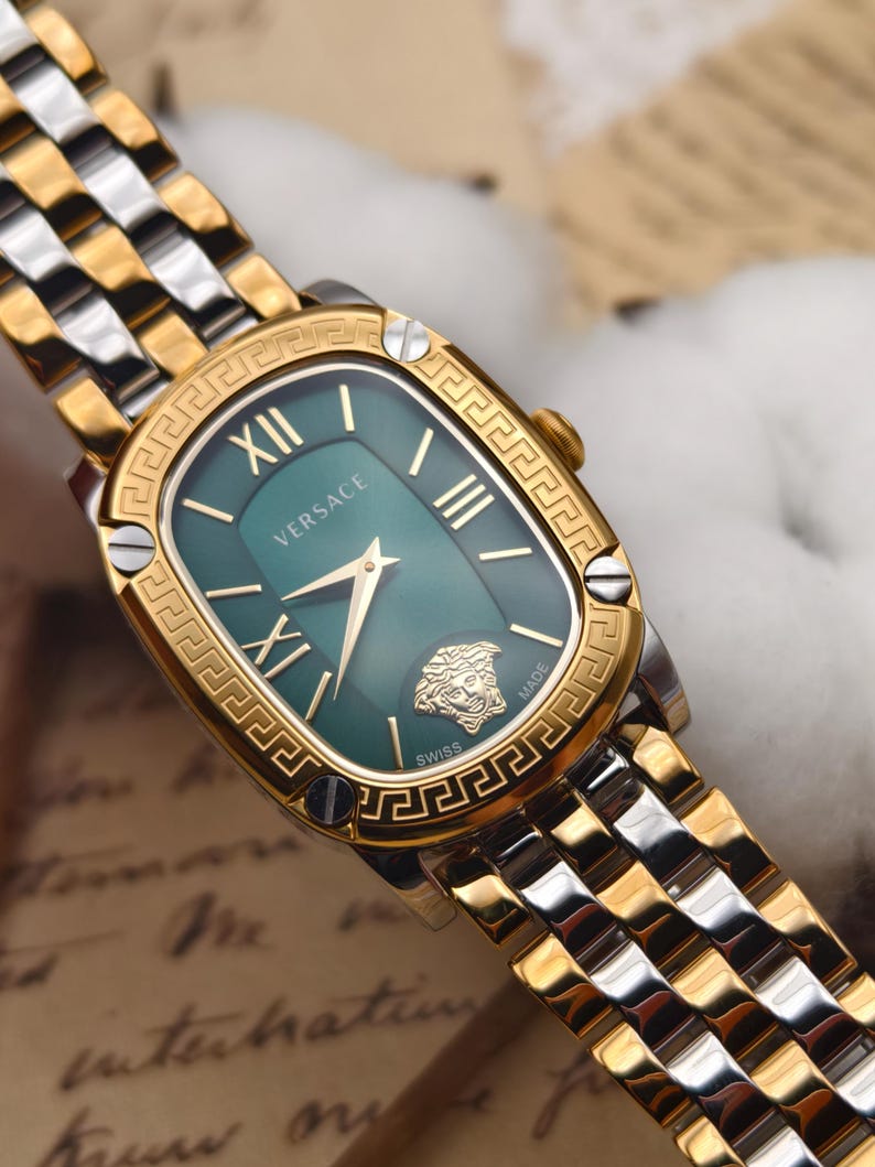 Puede incluir: Un reloj Versace dorado y plateado con esfera verde. El reloj tiene una esfera rectangular con n&uacute;meros romanos y agujas doradas. La correa del reloj es una combinaci&oacute;n de eslabones dorados y plateados. El logotipo de Versace y una cabeza de Medusa son visibles en la esfera.