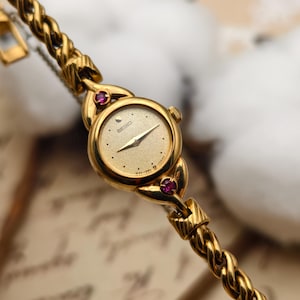 May include: A gold-tone Seiko wristwatch featuring a round, textured champagne dial with gold hands and markers. The watch case is accented with two small, round, deep pink stones at the top and bottom, connected to a twisted rope-style metal bracelet.