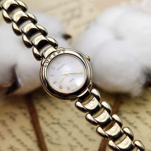 May include: A gold-tone Seiko Lucent wristwatch featuring a round white dial with gold Roman numeral hour markers and gold hands. The watch has a polished gold-tone metal link bracelet with a smooth, rounded design and a small crown on the side.