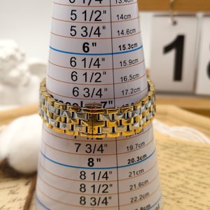 Puede incluir: Un brazalete dorado y plateado se mide en un medidor de brazalete blanco. El medidor tiene medidas en pulgadas y cent&iacute;metros, que van desde 12,7 cm hasta 22,8 cm. El brazalete es de estilo eslab&oacute;n.