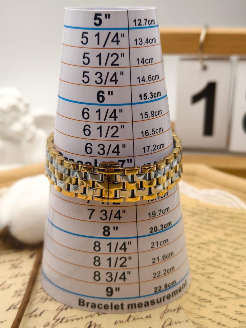 Puede incluir: Un brazalete dorado y plateado se mide en un medidor de brazalete blanco. El medidor tiene medidas en pulgadas y cent&iacute;metros, que van desde 12,7 cm hasta 22,8 cm. El brazalete es de estilo eslab&oacute;n.