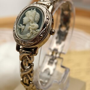 Puede incluir: Un reloj anal&oacute;gico de tono plateado con una esfera ovalada que presenta un perfil de camafeo blanco en relieve sobre un fondo gris oscuro. El reloj cuenta con una pulsera de eslabones met&aacute;licos texturizados y una corona en el lateral.