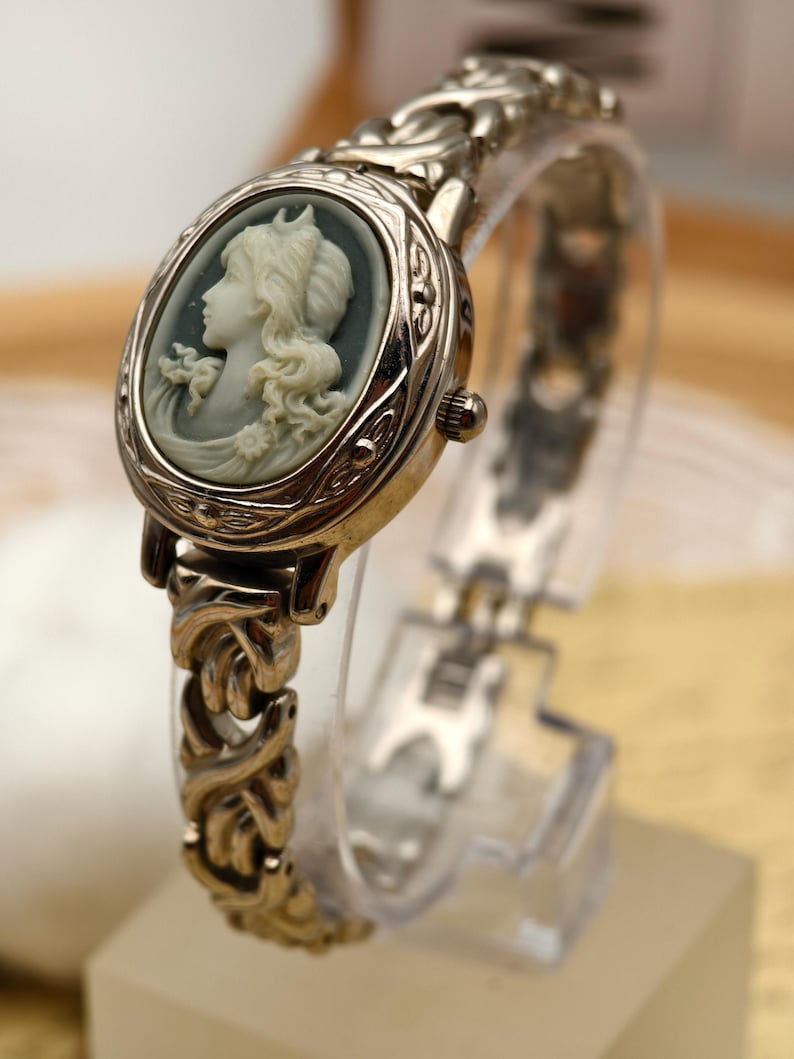 Puede incluir: Un reloj anal&oacute;gico de tono plateado con una esfera ovalada que presenta un perfil de camafeo blanco en relieve sobre un fondo gris oscuro. El reloj cuenta con una pulsera de eslabones met&aacute;licos texturizados y una corona en el lateral.