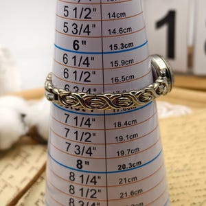 Puede incluir: Un reloj de metal plateado con una pulsera de eslabones entrelazados y texturizados. El reloj est&aacute; colocado sobre una herramienta de medici&oacute;n c&oacute;nica blanca, indicando un tama&ntilde;o de pulsera de unos 16,5 cm a 17 cm de circunferencia.