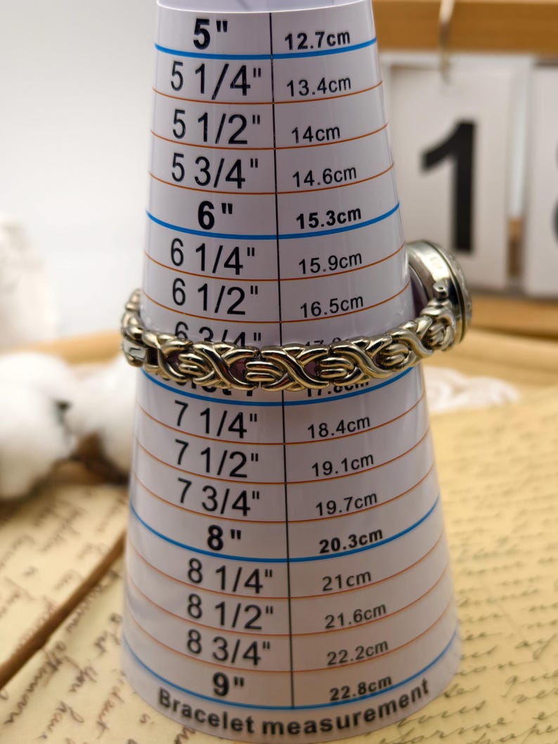 Puede incluir: Un reloj de metal plateado con una pulsera de eslabones entrelazados y texturizados. El reloj est&aacute; colocado sobre una herramienta de medici&oacute;n c&oacute;nica blanca, indicando un tama&ntilde;o de pulsera de unos 16,5 cm a 17 cm de circunferencia.